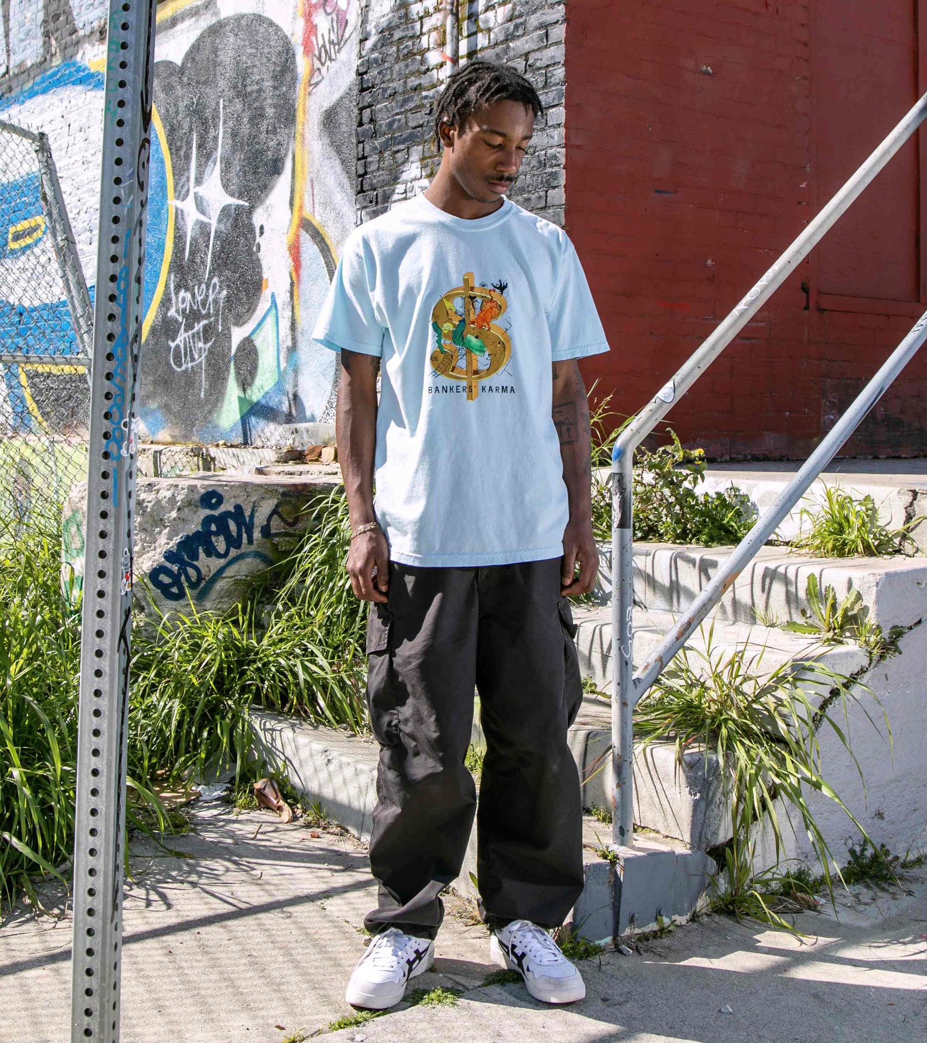 Wide shot of male model outside wearing chambray blue colored unisex cotton graphic t-shirt with Bankers Karma print visible.