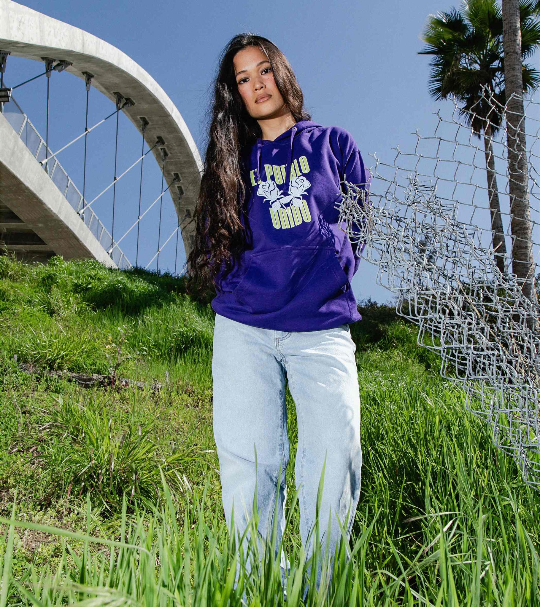 Wide shot of woman standing in a field wearing El Pueblo Unido classic fit midweight purple hoodie with front graphic