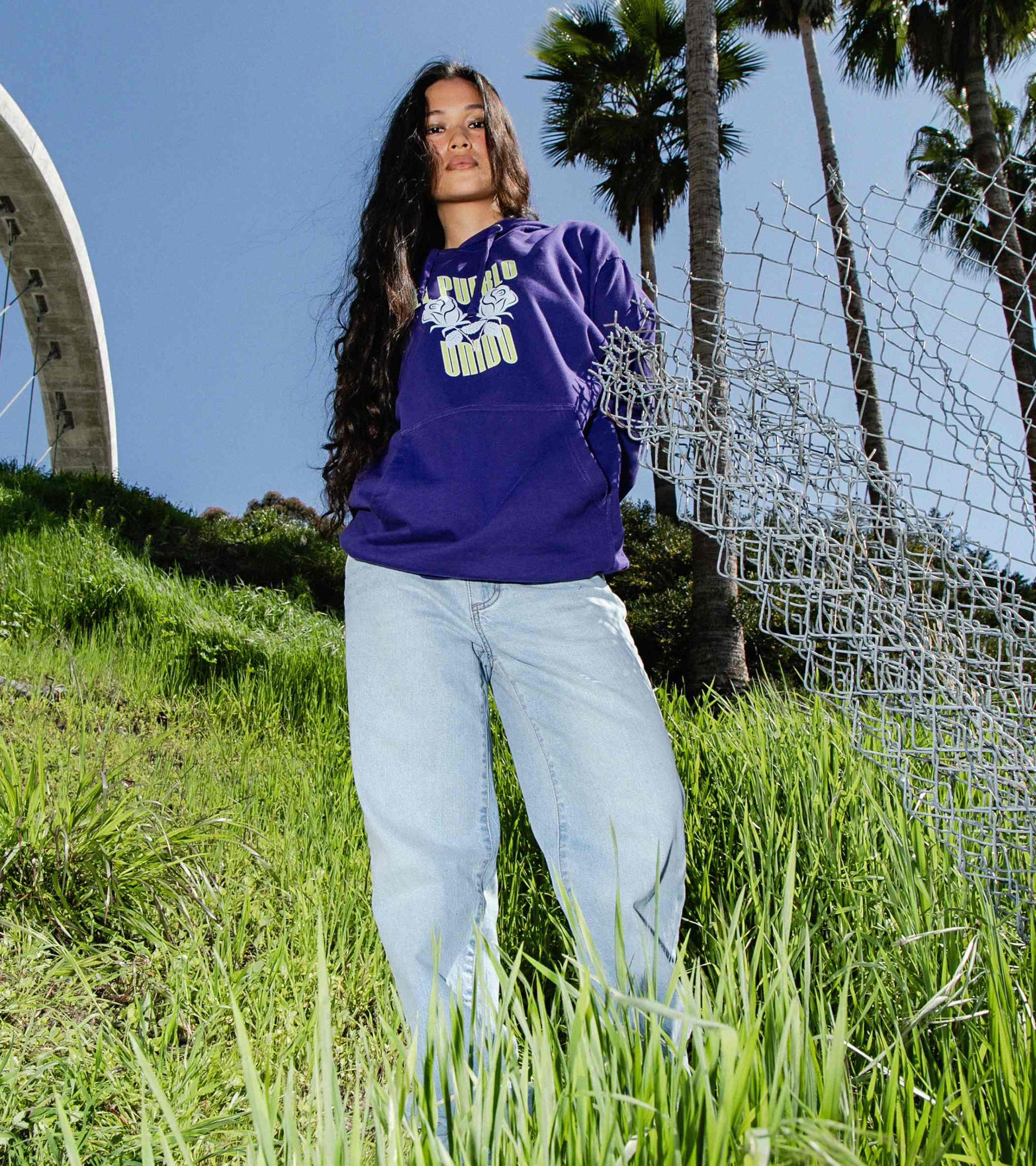 Wide shot of woman wearing classic fit midweight purple hoodie with yellow and white front graphic that says El Pueblo Unido 