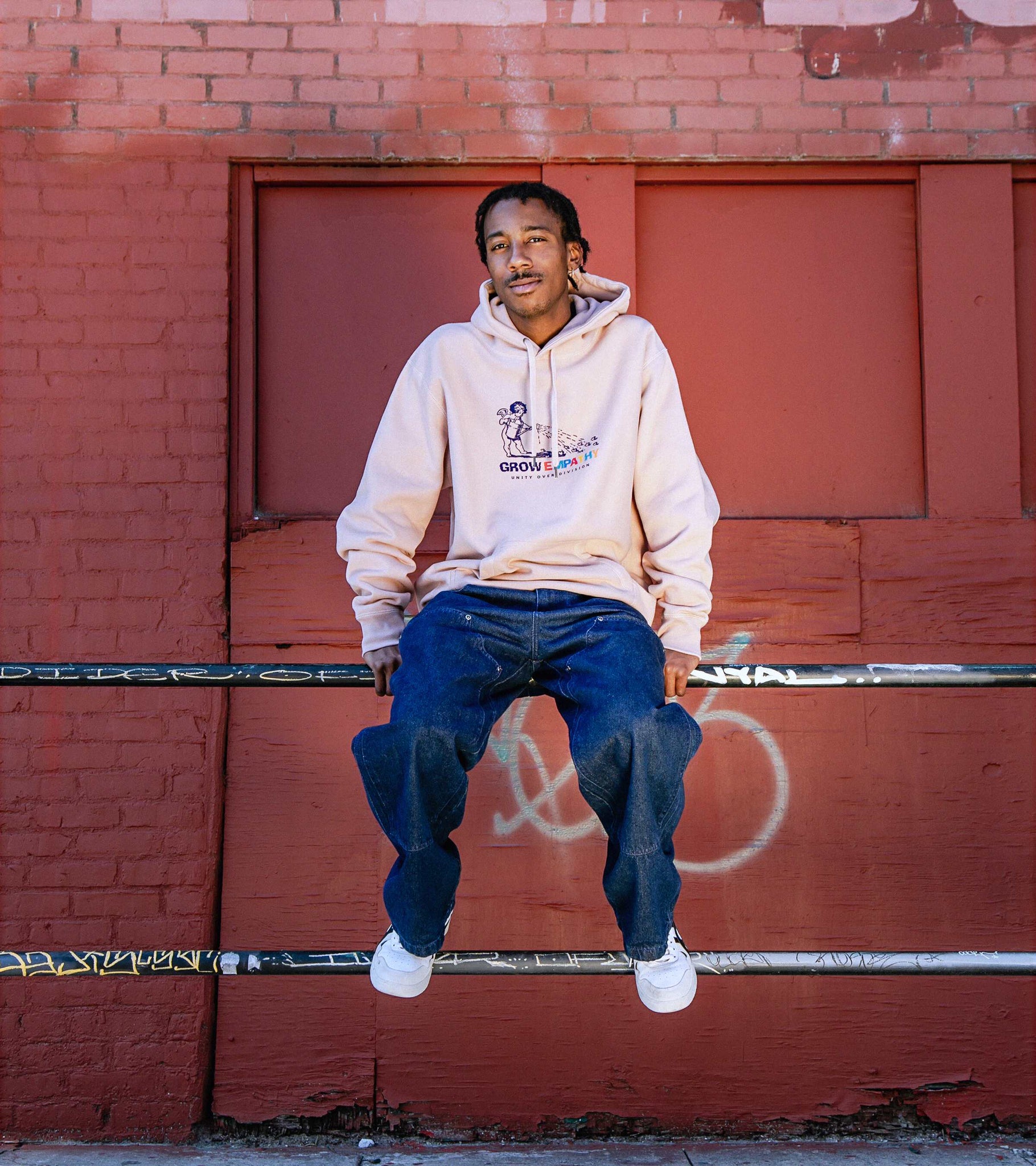 Wide shot of male model sitting on rail wearing Grow Empathy Unity Over Division heavyweight cross-grain hoodie in dusty pink