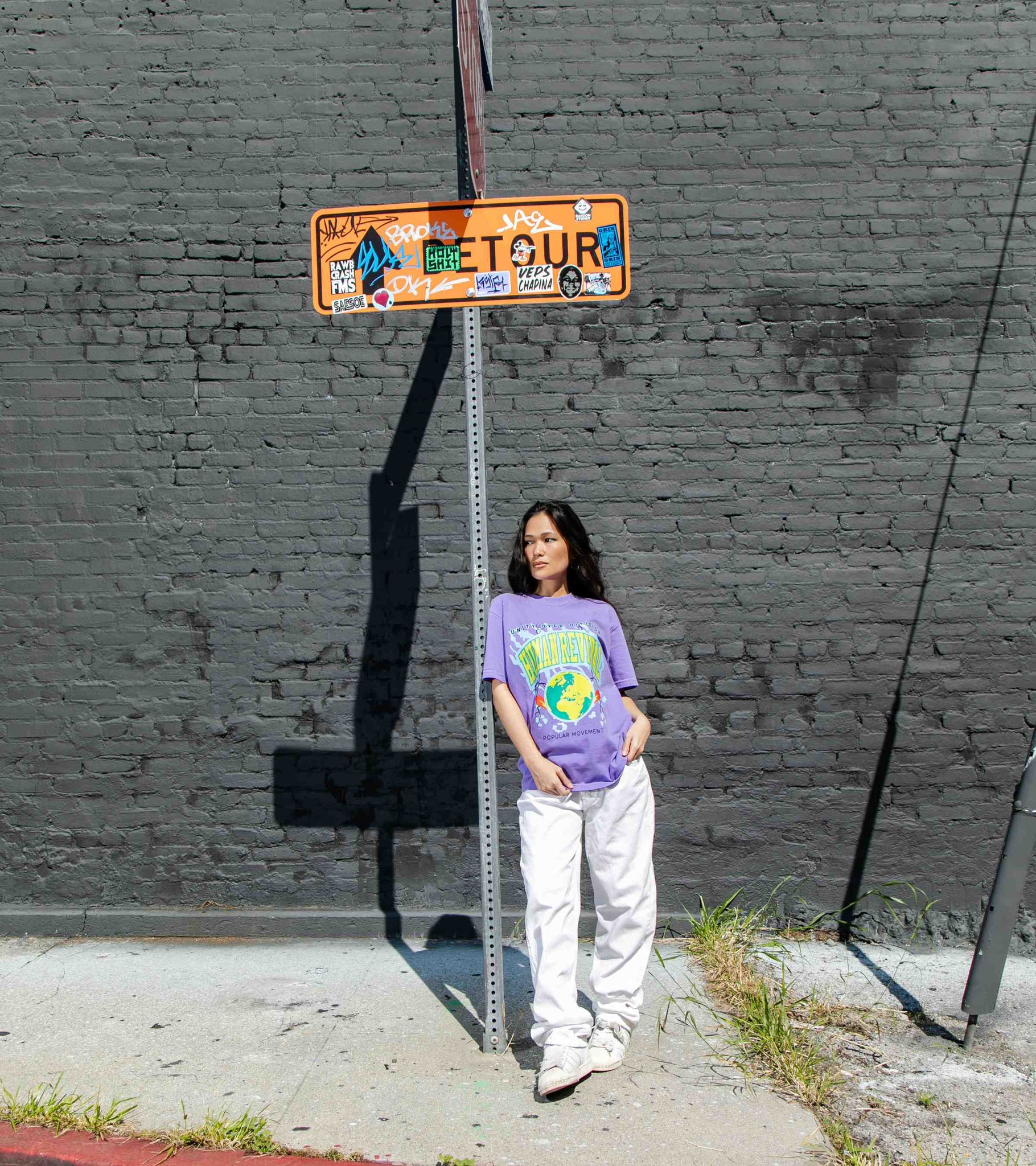 Wide shot of woman model standing by a street sign wearing summery violet colored unisex cotton graphic t-shirt