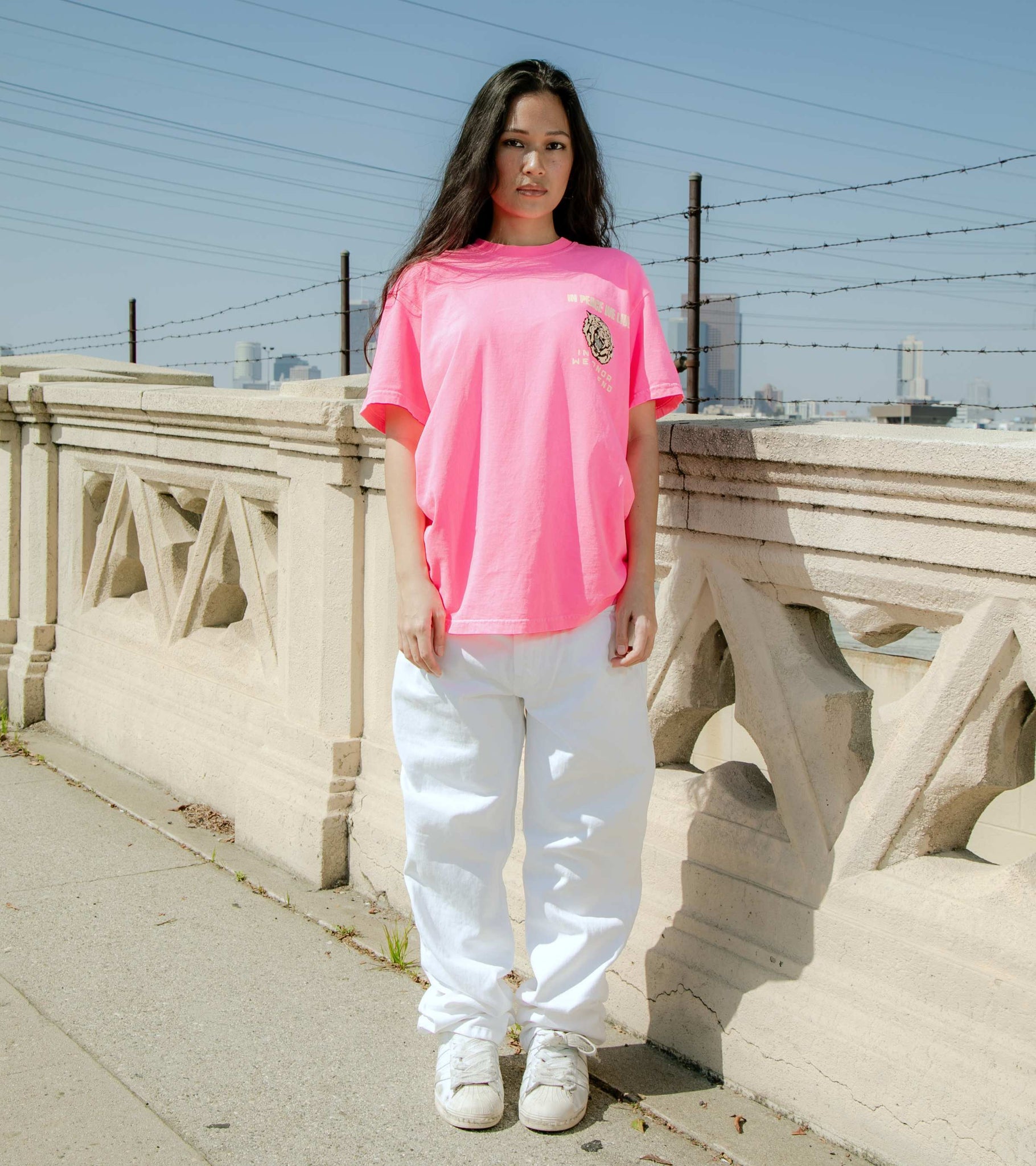 Full body shot of female model wearing graphic t-shirt in neon pink with In Peace We Live In Honor We Defend front print.