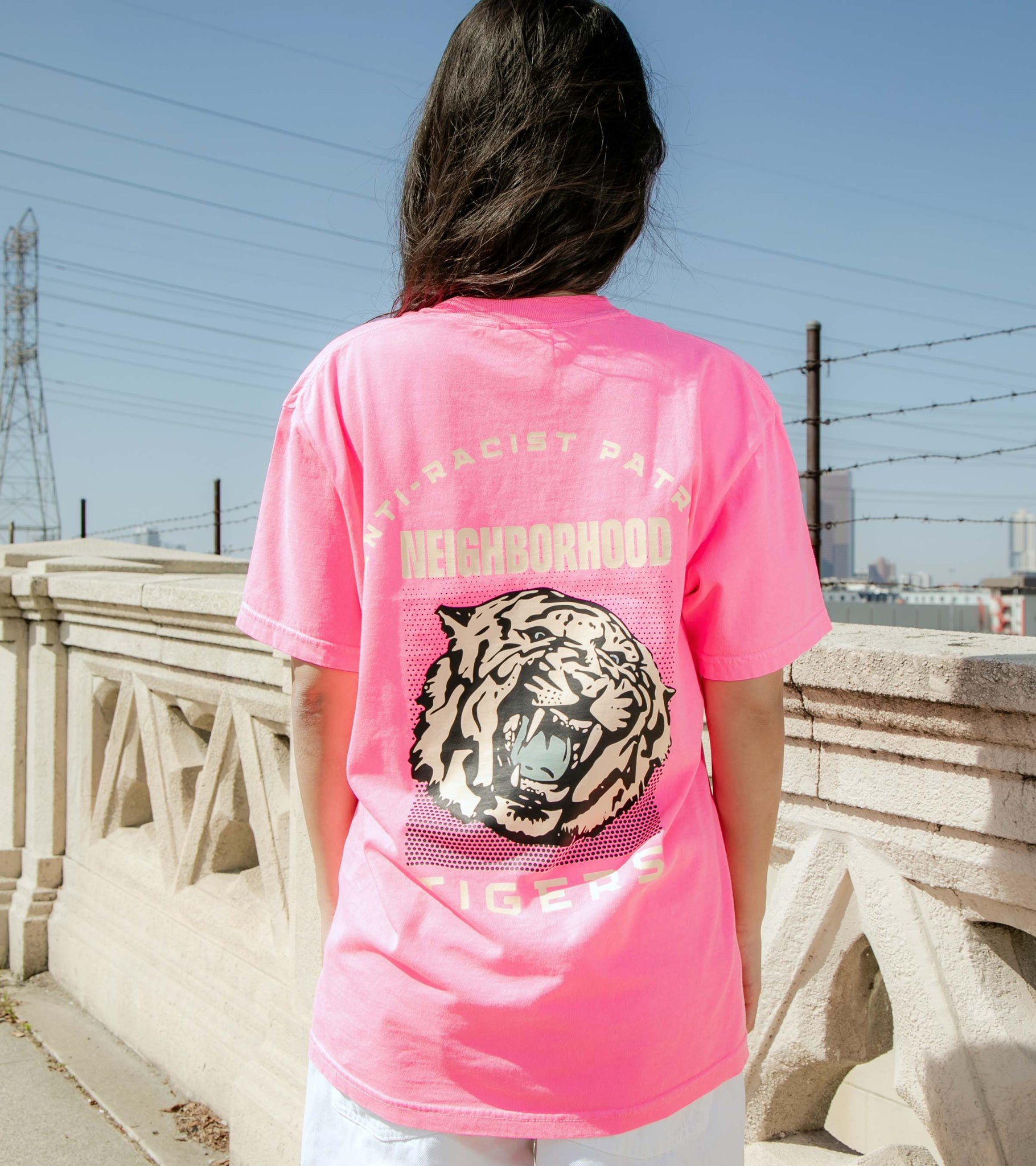 Medium shot of female model in neon pink graphic t-shirt with Anti-Racist Patrol, Neighborhood Tigers visible back print.