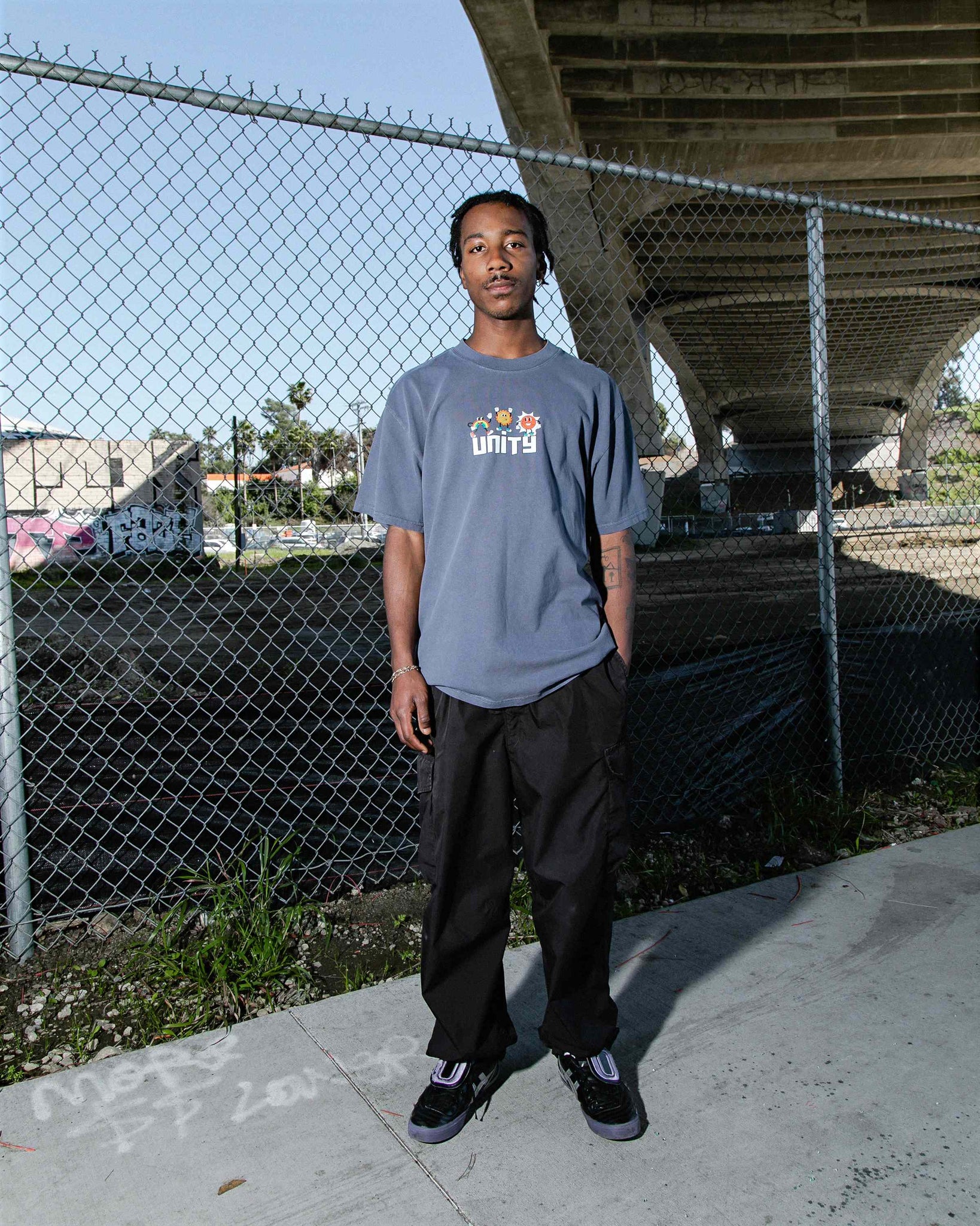 Wide shot of male model standing in front of fence wearing Unity faded indigo oversized fit unisex cotton graphic t-shirt